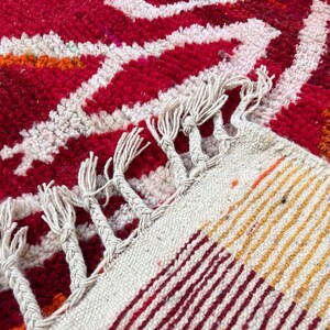 May include: Close-up of a vibrant, hand-woven rug with a bold geometric pattern. The rug features a rich red background with white, orange, and yellow accents. The edges are finished with a fringe.