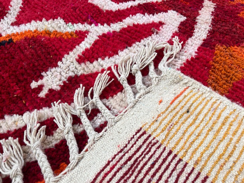 May include: Close-up of a vibrant, hand-woven rug with a bold geometric pattern. The rug features a rich red background with white, orange, and yellow accents. The edges are finished with a fringe.