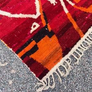 May include: Close-up of a vibrant, hand-woven rug with a geometric design. The rug features a rich color palette of red, orange, black, and white. The edges are finished with a fringe.