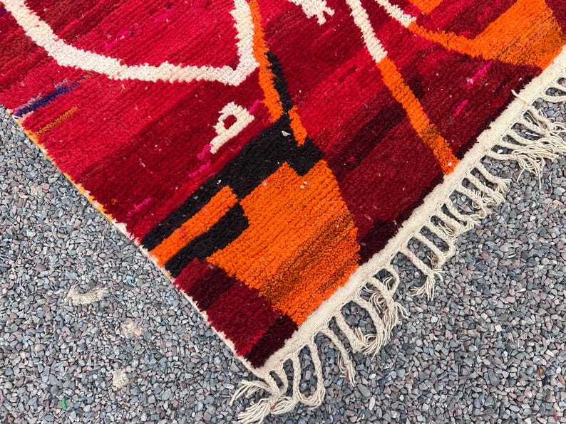 May include: Close-up of a vibrant, hand-woven rug with a geometric design. The rug features a rich color palette of red, orange, black, and white. The edges are finished with a fringe.