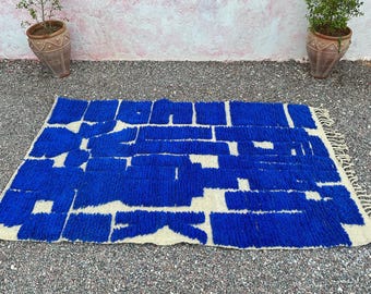 May include: A rectangular rug with a blue and white geometric pattern. The rug has a thick pile and a fringe on one end. The design features blue rectangular shapes on a white background. The rug is placed on a gravel surface.