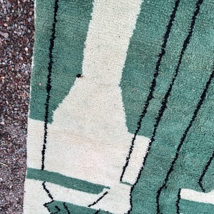 May include: Close-up of a green and white geometric patterned rug. The rug features a design of green rectangles and white shapes, with black lines. The rug is made of a soft, textured material.