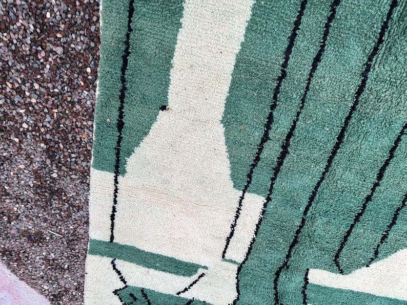 May include: Close-up of a green and white geometric patterned rug. The rug features a design of green rectangles and white shapes, with black lines. The rug is made of a soft, textured material.