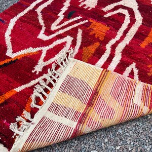 May include: A vibrant red rug with a geometric pattern in white, orange, and black. The rug's design features abstract shapes and lines, with a fringed edge. The underside of the rug displays a striped pattern in shades of red, yellow, and cream.