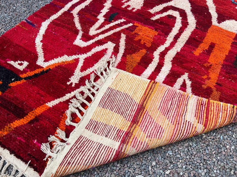 May include: A vibrant red rug with a geometric pattern in white, orange, and black. The rug's design features abstract shapes and lines, with a fringed edge. The underside of the rug displays a striped pattern in shades of red, yellow, and cream.