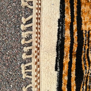 May include: Close-up of a textured rug with a cream-colored central panel bordered by brown stitching and tassels. The right side features black and orange vertical stripes, creating a bold contrast. The rug's design suggests a bohemian or tribal aesthetic.