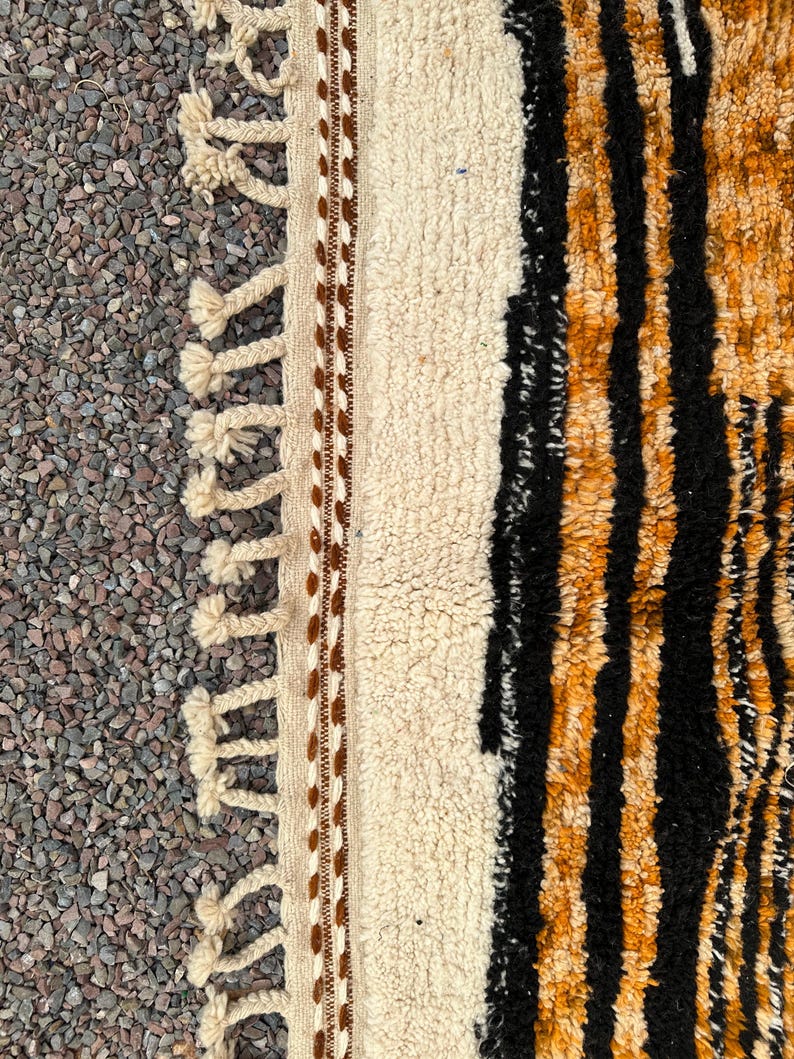 May include: Close-up of a textured rug with a cream-colored central panel bordered by brown stitching and tassels. The right side features black and orange vertical stripes, creating a bold contrast. The rug's design suggests a bohemian or tribal aesthetic.