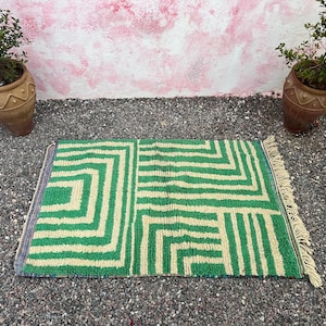 Pode incluir: Um tapete retangular com padr&atilde;o geom&eacute;trico verde e bege com borda com franjas. O tapete &eacute; colocado sobre uma superf&iacute;cie de cascalho, com duas plantas em vasos ao fundo. O design do tapete apresenta uma s&eacute;rie de formas angulares.