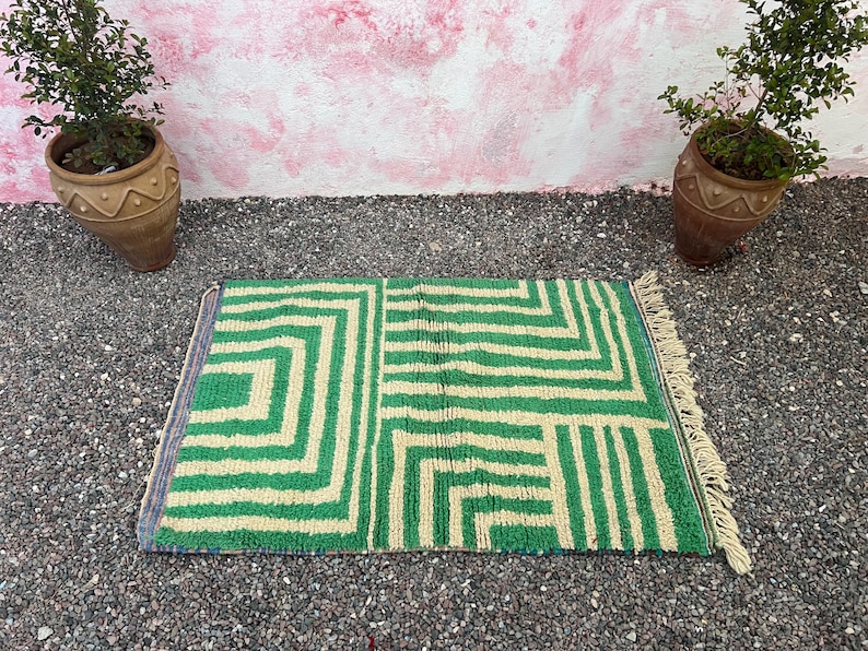 Pode incluir: Um tapete retangular com padr&atilde;o geom&eacute;trico verde e bege com borda com franjas. O tapete &eacute; colocado sobre uma superf&iacute;cie de cascalho, com duas plantas em vasos ao fundo. O design do tapete apresenta uma s&eacute;rie de formas angulares.