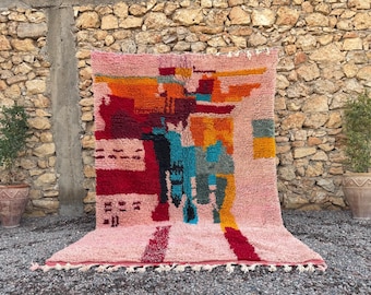 May include: A pink, abstract design rug with a textured surface. The rug features a mix of colors including red, orange, blue, and yellow. The rug has a fringe along the bottom edge and is displayed against a stone wall.
