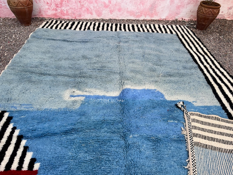 May include: A large, rectangular rug with a blue and white design. The rug has a textured surface and a border of black and white stripes. The rug's design includes a lighter blue shape in the center.