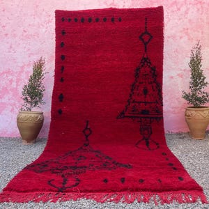 May include: A red Moroccan rug with black geometric designs. The rug has a fringe border and features a large tree-like pattern in the center.
