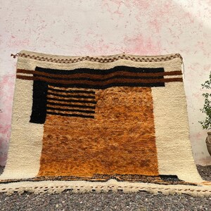 May include: A rectangular, hand-woven rug with a geometric design. The rug features a cream-colored background with brown, black, and orange geometric shapes. The rug is displayed in front of a pink wall, with potted plants on either side.