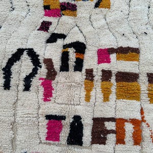 May include: Cream-colored, textured rug with a geometric pattern. The design incorporates shapes in black, brown, pink, orange, and yellow. The rug's surface has a plush, woven texture, creating a visually interesting and tactile appeal.