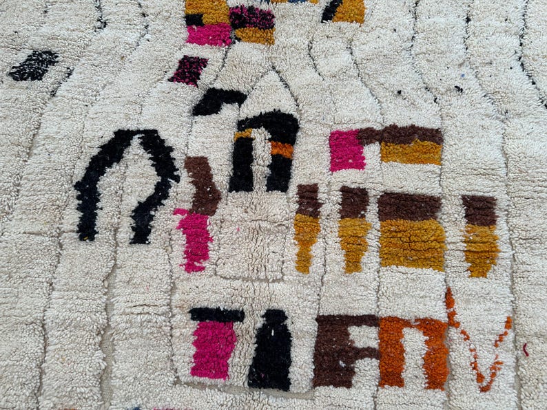 May include: Cream-colored, textured rug with a geometric pattern. The design incorporates shapes in black, brown, pink, orange, and yellow. The rug's surface has a plush, woven texture, creating a visually interesting and tactile appeal.