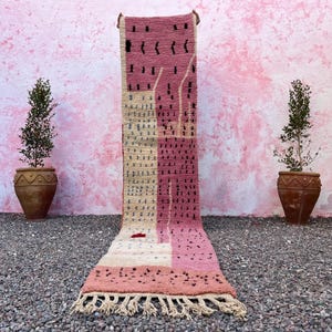 May include: A long, rectangular rug with a pink, beige, and white geometric pattern. The rug has a fringe on the bottom edge and is displayed against a pink wall. Two potted plants flank the rug.