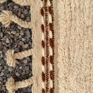 May include: Close-up of a beige and brown woven rug with a textured, shaggy pile. The rug features a braided border with tassels and a central stripe with brown and white decorative elements. The rug is placed on a bed of small, gray stones.