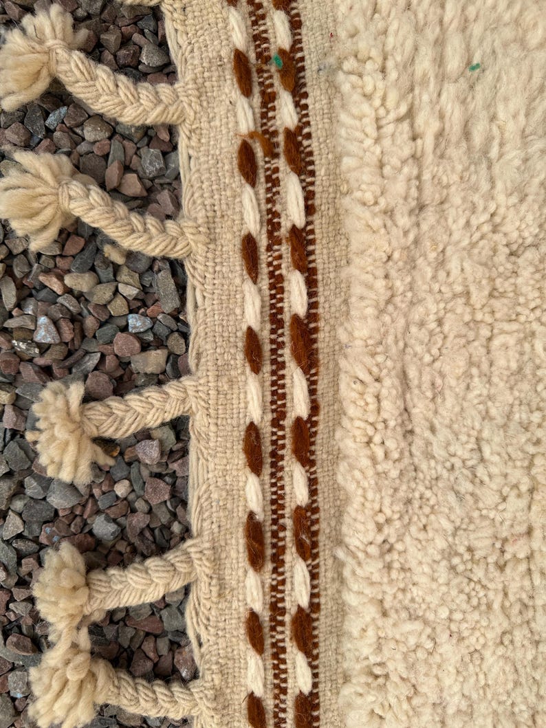 May include: Close-up of a beige and brown woven rug with a textured, shaggy pile. The rug features a braided border with tassels and a central stripe with brown and white decorative elements. The rug is placed on a bed of small, gray stones.