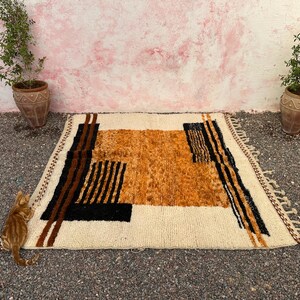 May include: A rectangular, cream-colored rug with an abstract design in shades of brown, black, and orange. The rug features geometric shapes and vertical stripes. The rug has a fringed edge and is placed on a textured surface.