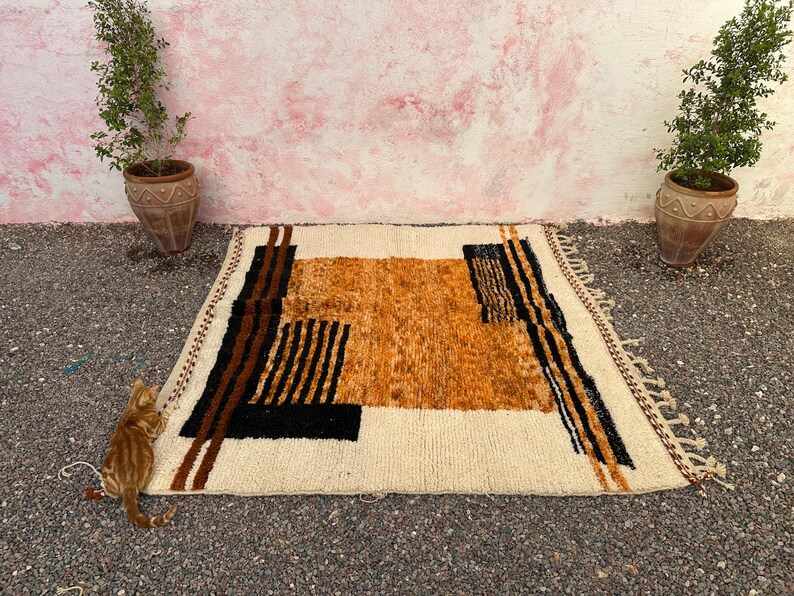 May include: A rectangular, cream-colored rug with an abstract design in shades of brown, black, and orange. The rug features geometric shapes and vertical stripes. The rug has a fringed edge and is placed on a textured surface.
