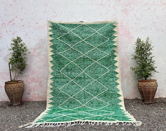 May include: A rectangular green rug with a diamond pattern and cream-colored zigzag edges. The rug has a fringe at the bottom and is displayed against a pink and white wall. Two potted plants flank the rug.