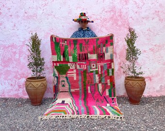 May include: A pink and green Moroccan rug with geometric patterns. The rug is being held up by a person wearing a colorful hat.