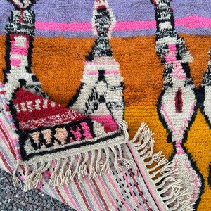 May include: A colorful, textured rug with a geometric pattern. The rug features a combination of orange, yellow, purple, and pink hues. The design includes diamond and vertical shapes with fringe detailing on the edge. The rug is made of a thick, woven material.