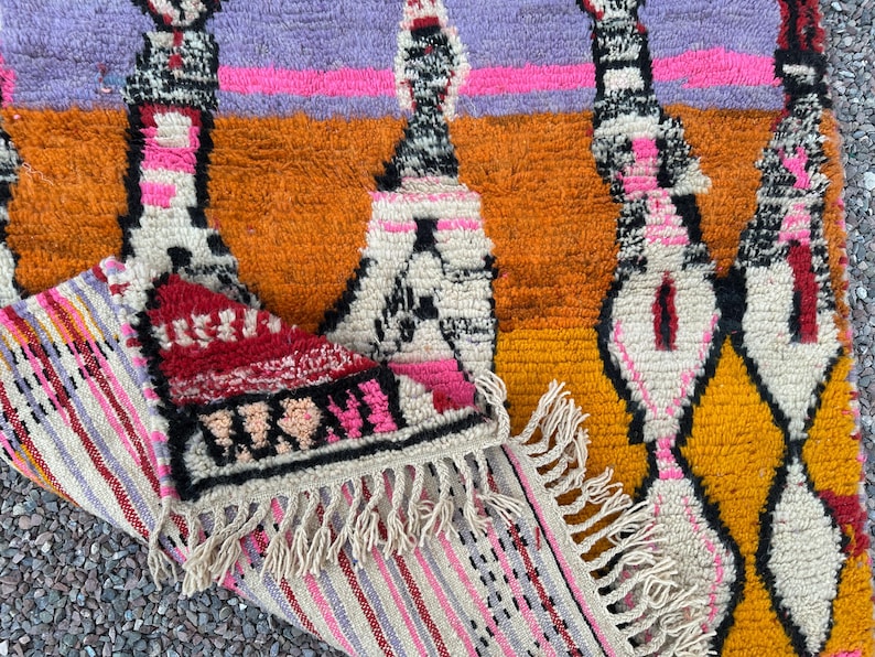 May include: A colorful, textured rug with a geometric pattern. The rug features a combination of orange, yellow, purple, and pink hues. The design includes diamond and vertical shapes with fringe detailing on the edge. The rug is made of a thick, woven material.