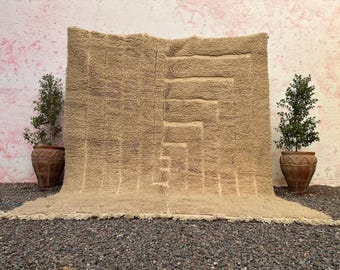 Op de afbeelding: Een groot, beige tapijt met een geometrisch patroon wordt getoond tegen een roze en witte muur. Het tapijt heeft een getextureerd oppervlak en een franje langs de onderrand. Twee potplanten flankeren het tapijt en voegen groen toe.
