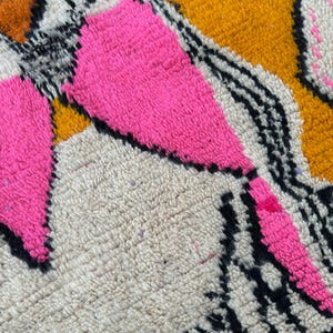 May include: Close-up of a colorful rug with a geometric design. The rug features bright pink, orange, black, and white patterns. The texture is thick and woven, with a soft, plush feel.