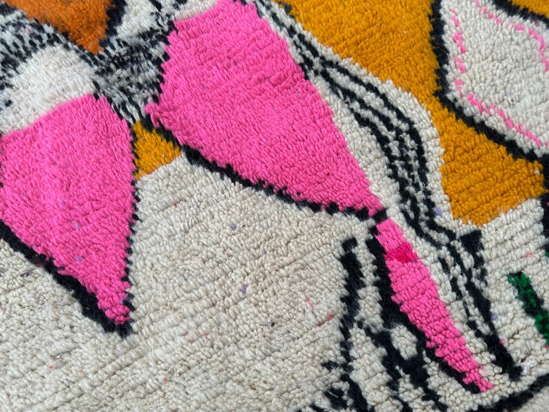 May include: Close-up of a colorful rug with a geometric design. The rug features bright pink, orange, black, and white patterns. The texture is thick and woven, with a soft, plush feel.