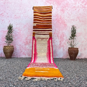 Op de afbeelding: Een lang, smal tapijt met een witte achtergrond en kleurrijke strepen in tinten oranje, roze en rood. Het tapijt heeft aan beide uiteinden franjes.