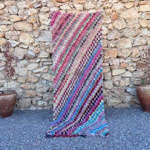 May include: A rectangular rug featuring a vibrant, multi-colored pattern of diagonal stripes. The textile is composed of various woven fabric strips in shades of pink, red, blue, purple, and teal, creating a textured, shaggy appearance.