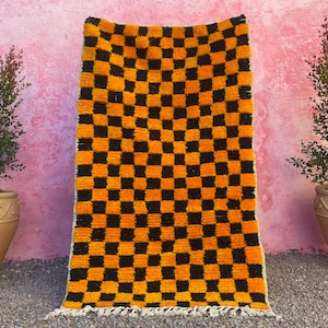 May include: A black and orange checkered rug with a fringe border. The rug is made of a soft, fluffy material and has a geometric pattern.