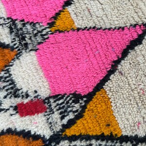 May include: Close-up of a colorful rug with a geometric pattern. The rug features bright pink, orange, yellow, black, and white sections. The texture appears soft and plush, with a high pile.