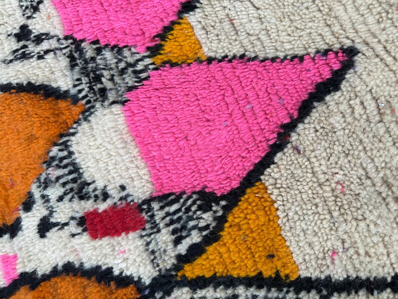May include: Close-up of a colorful rug with a geometric pattern. The rug features bright pink, orange, yellow, black, and white sections. The texture appears soft and plush, with a high pile.