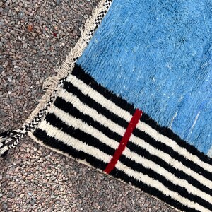 May include: A close-up of a blue rug with a black and white striped border. A thin red stripe runs vertically through the black and white stripes. The rug has a textured appearance and a fringed edge.