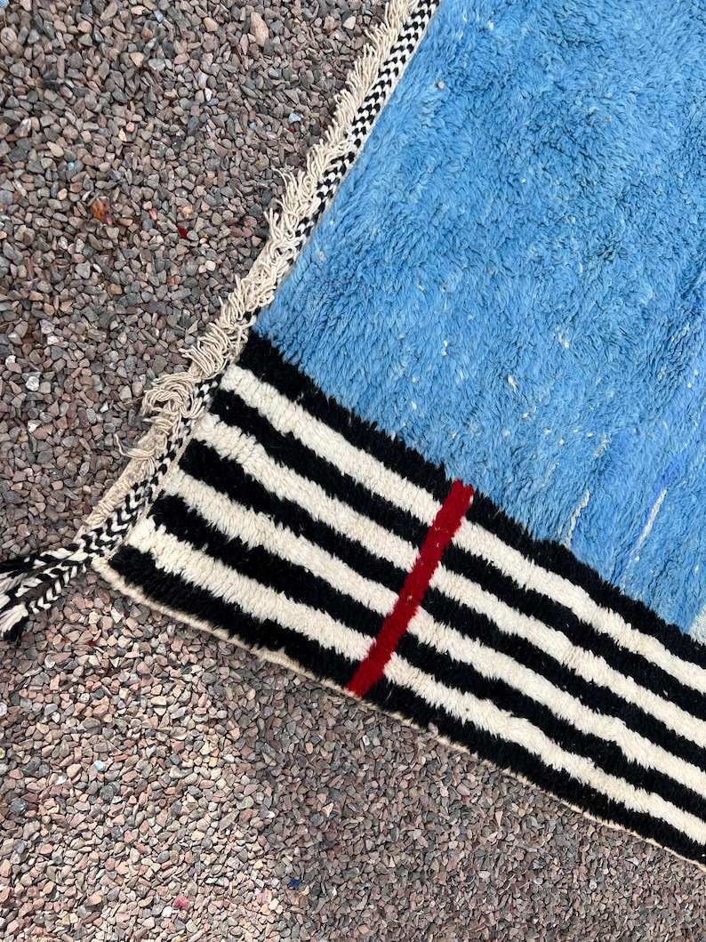 May include: A close-up of a blue rug with a black and white striped border. A thin red stripe runs vertically through the black and white stripes. The rug has a textured appearance and a fringed edge.