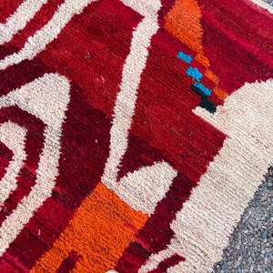 May include: Close-up of a vibrant, textured rug with a geometric design. The rug features a bold color palette of red, orange, and cream, with accents of blue and black. The design includes abstract shapes and lines.