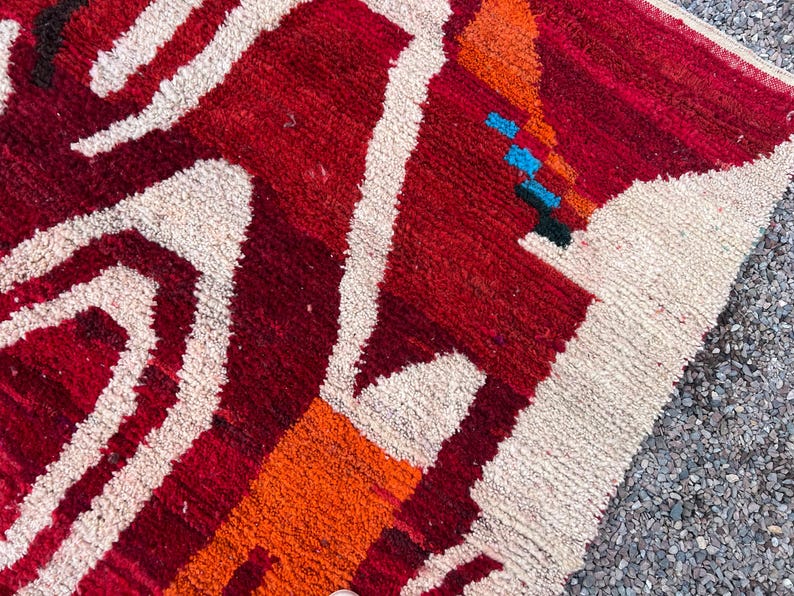 May include: Close-up of a vibrant, textured rug with a geometric design. The rug features a bold color palette of red, orange, and cream, with accents of blue and black. The design includes abstract shapes and lines.