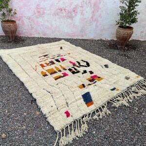 May include: Cream-colored, rectangular rug with a geometric design in shades of orange, pink, blue, and black. The rug has a fringed edge and a textured surface, suitable for a living room or bedroom.