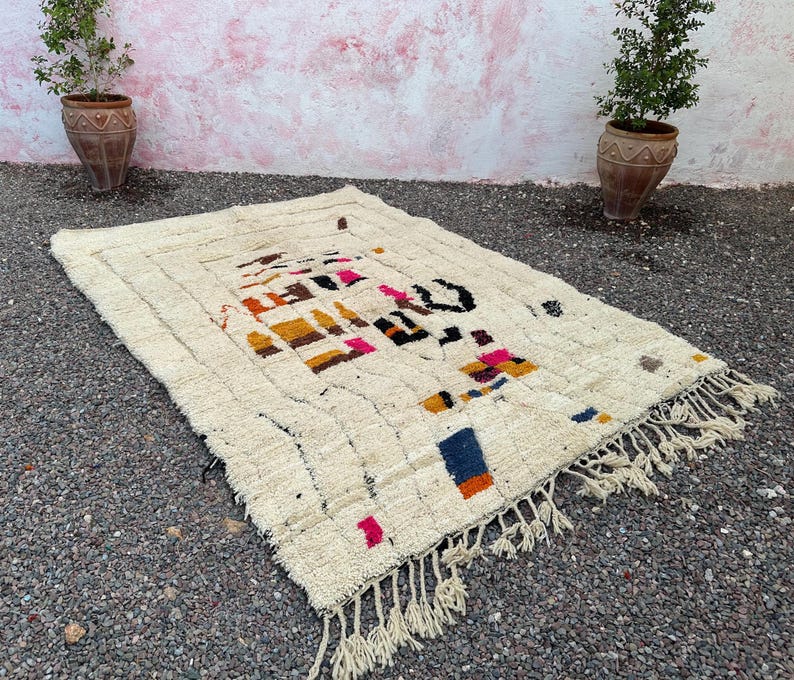 May include: Cream-colored, rectangular rug with a geometric design in shades of orange, pink, blue, and black. The rug has a fringed edge and a textured surface, suitable for a living room or bedroom.