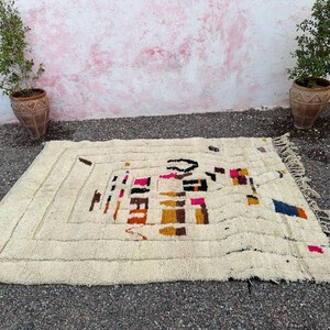 May include: Cream-colored rectangular rug with a geometric design in black, pink, orange, and blue. The rug has a textured surface and a fringe along one edge. The rug is placed on a dark surface with plants in terracotta pots on either side.