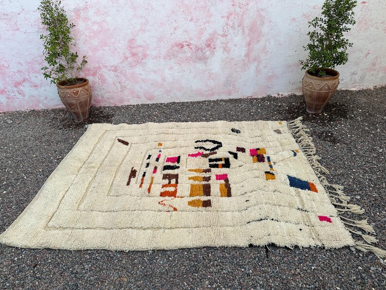 May include: Cream-colored rectangular rug with a geometric design in black, pink, orange, and blue. The rug has a textured surface and a fringe along one edge. The rug is placed on a dark surface with plants in terracotta pots on either side.