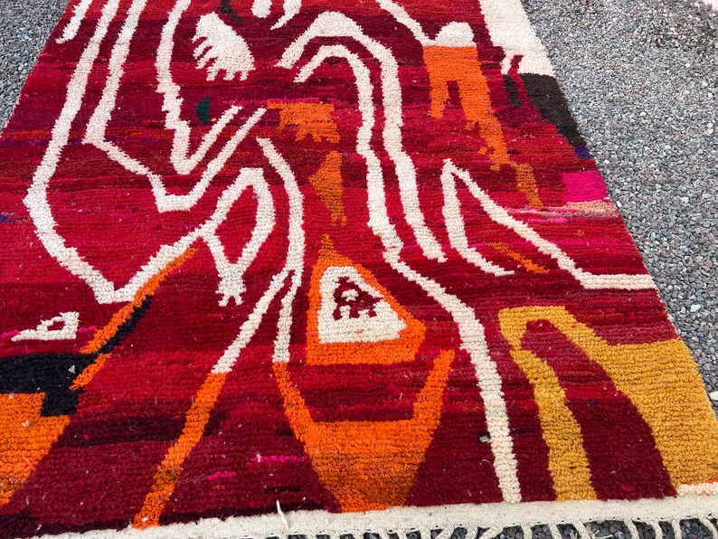 May include: A vibrant, hand-woven rug with a geometric design. The rug features a rich red background with white, orange, and yellow abstract patterns. The edges are finished with a white fringe. This rug is suitable for home decor.