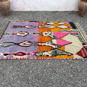May include: A rectangular, hand-woven rug with a vibrant geometric design. The rug features sections of purple, orange, pink, and yellow, with black and white accents. It has a fringed edge and is likely made of wool.