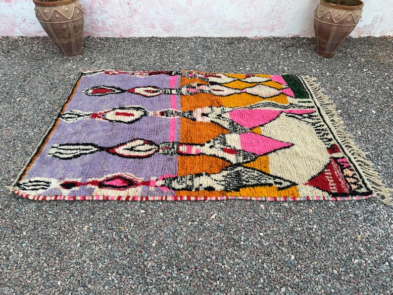 May include: A rectangular, hand-woven rug with a vibrant geometric design. The rug features sections of purple, orange, pink, and yellow, with black and white accents. It has a fringed edge and is likely made of wool.
