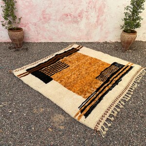 May include: A square, cream-colored rug with a geometric design. The rug features a central orange-brown section, bordered by black and brown stripes and accents. The edges have a fringe detail. The rug is placed on a gravel surface.