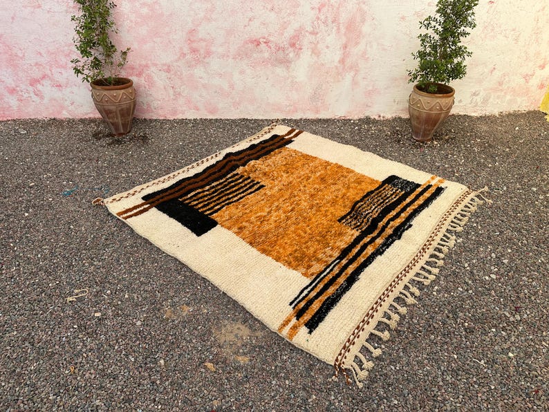 May include: A square, cream-colored rug with a geometric design. The rug features a central orange-brown section, bordered by black and brown stripes and accents. The edges have a fringe detail. The rug is placed on a gravel surface.