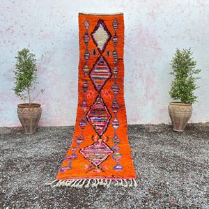 Peut inclure: Un tapis de couloir orange vif avec un motif de losanges. Le tapis présente un fond orange vif avec des formes de losanges colorées et une frange en bas. Le tapis est exposé sur une surface de gravier.